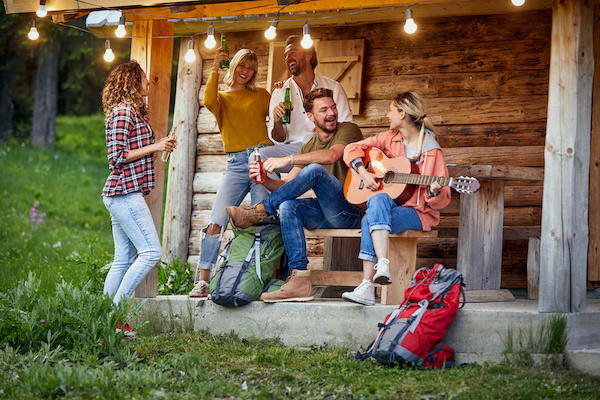 Young cheerful friends toasting and drinking beer in front of wooden cottage on the terrace.  Girl playing guitar. Summertime garden celebration and fun. Friends, togetherness, fun, joy, celebration concept.